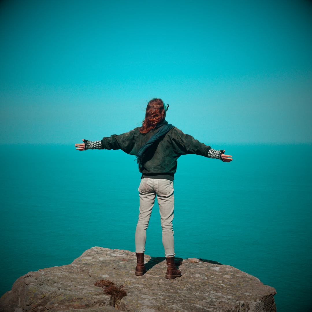 Ready to Thrive - Woman on top of rock cliff, arms open wide, looking out over the ocean